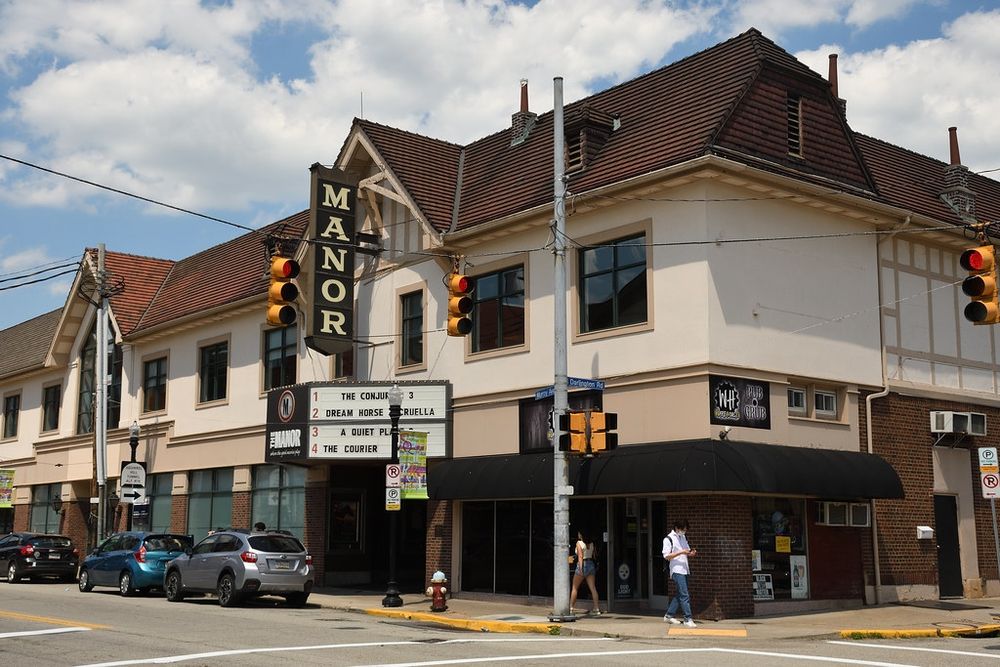 Photo of a movie theater from the street. A sign in front reads "Manor." The theater is at an intersection with a few parked cars and pedestrians.