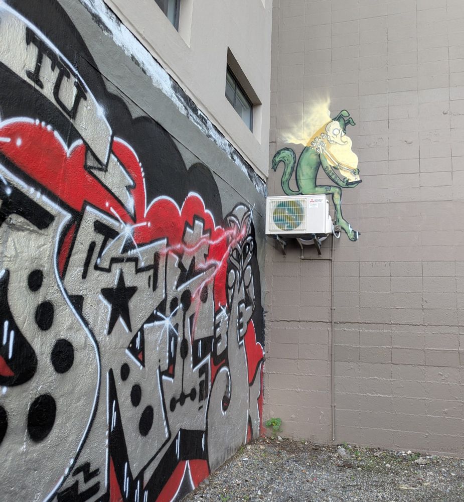 Graffiti of a little green something sitting on an HVAC unit on the side of a wall, reading something that strips the skin off his bones 