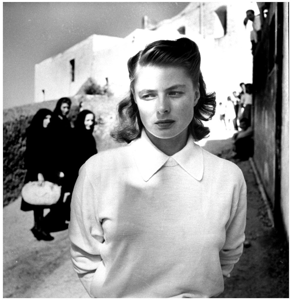 Black and white photograph the actress in a white blouse and sweater stands with her face half and sunlight half and Shadow looking off to the side a thoughtful or sad expression on her face over her shoulder we see three women dressed all in black looking in her Direction