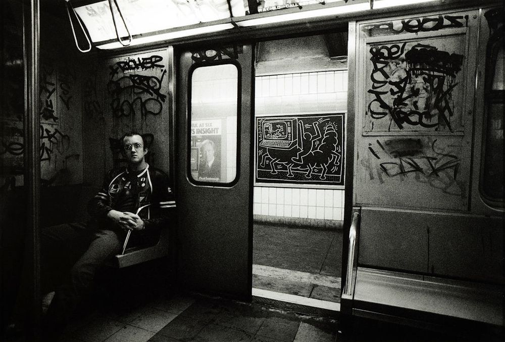 Black and white photograph of the artist sitting in subway car the interior of the subway is covered in graffiti just you know scribbles unreadable tags or at least unreadable by me through the half open door of the subway car we see on the station wall one of herrings on graffiti pieces as a poster