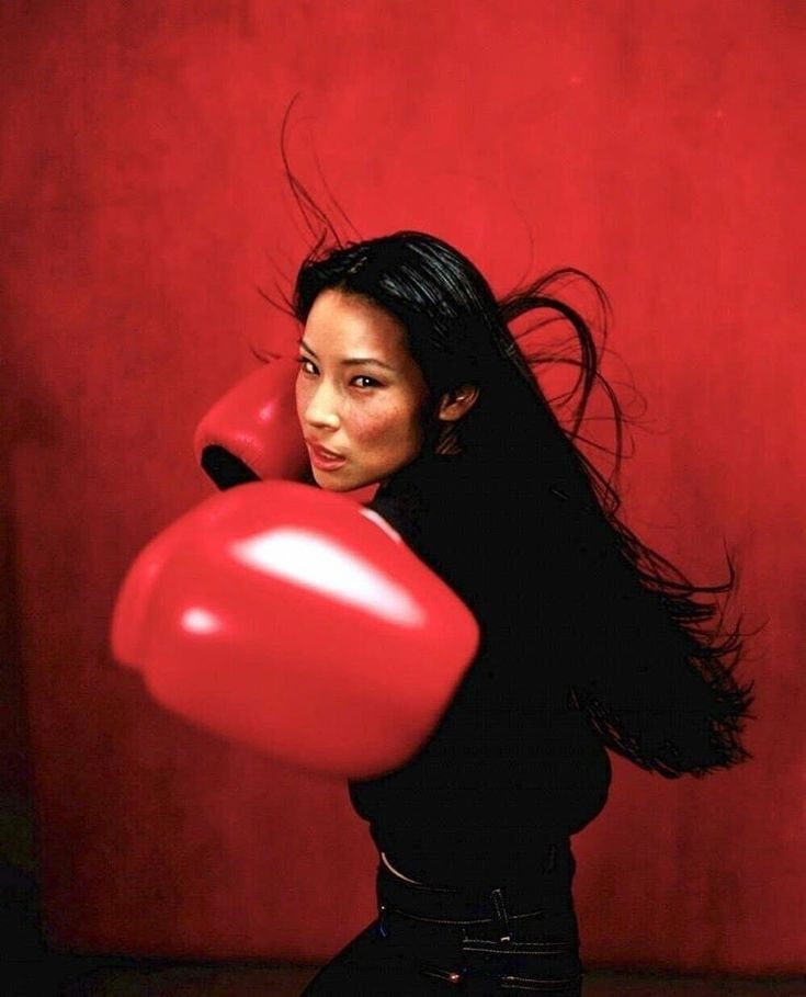 Photograph of the actress dressed in black wearing huge oversized red boxing gloves and standing in front of a red background his aggressive looking her face and has her right hand pulled back for a punch and her left hand up in guard 