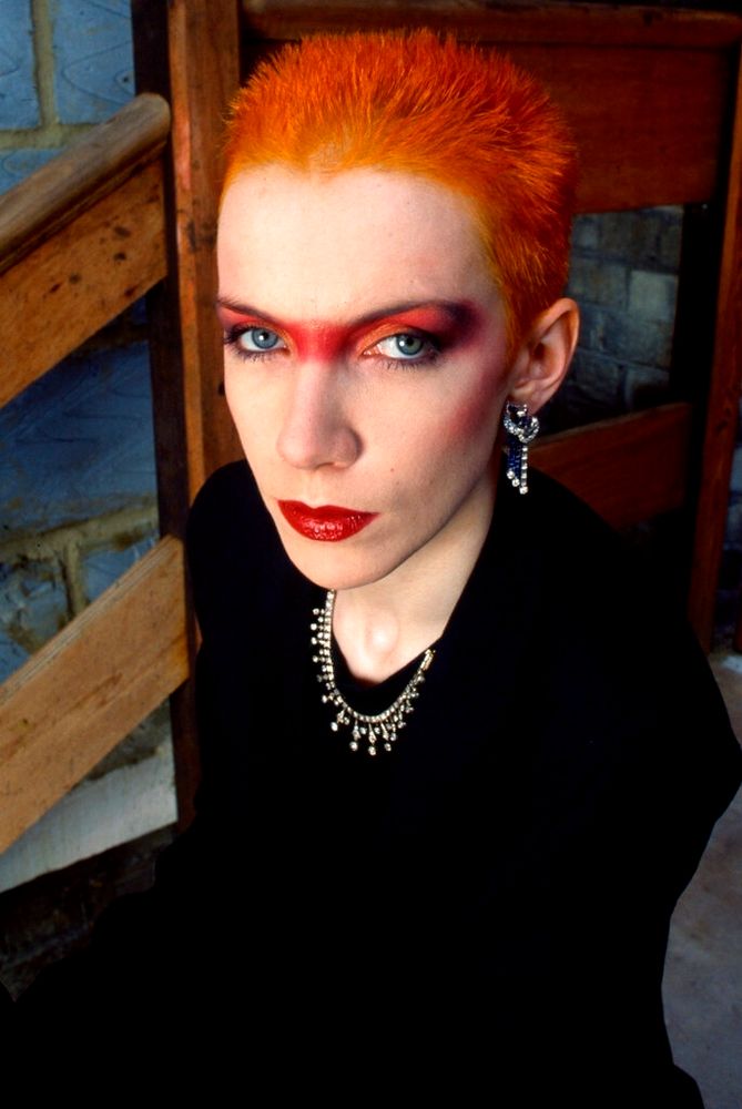 Photographed the center red orange hair red eyeshadow running in a stripe across the bridge of her nose very red lipstick black top diamond jewelry 