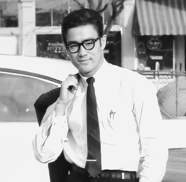 b&w photo of Bruce with a nerd look. He's wearing a skinny tie, with his jacket draped over his shoulder. He's wearing Clark Kent glasses, which is appropriate for a superman.