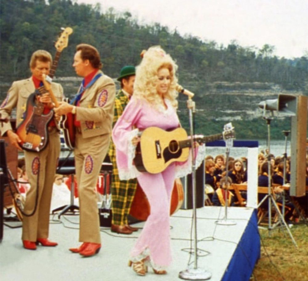 Dolly on stage c. early 1970s. Huge hair and a pink jumpsuit. I think the man right behind her in a Nudie suit is Porter Wagoner.