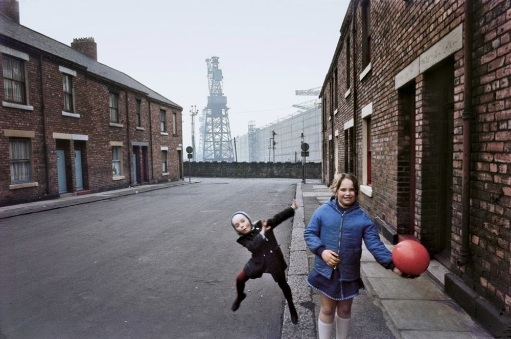 Photograph of a working class Street in the north of England down past the industry we can see what I think are docks industrial area with a high crane in between plain brick buildings there are two children on the sidewalk in the foreground a girl in a blue jacket with a big red ball and a very uncertain smile on her face behind her another child caught in mid dance or something and their face looking honestly they look like they could be some sort of cybernetic alien villain from Doctor Who an uncanny child