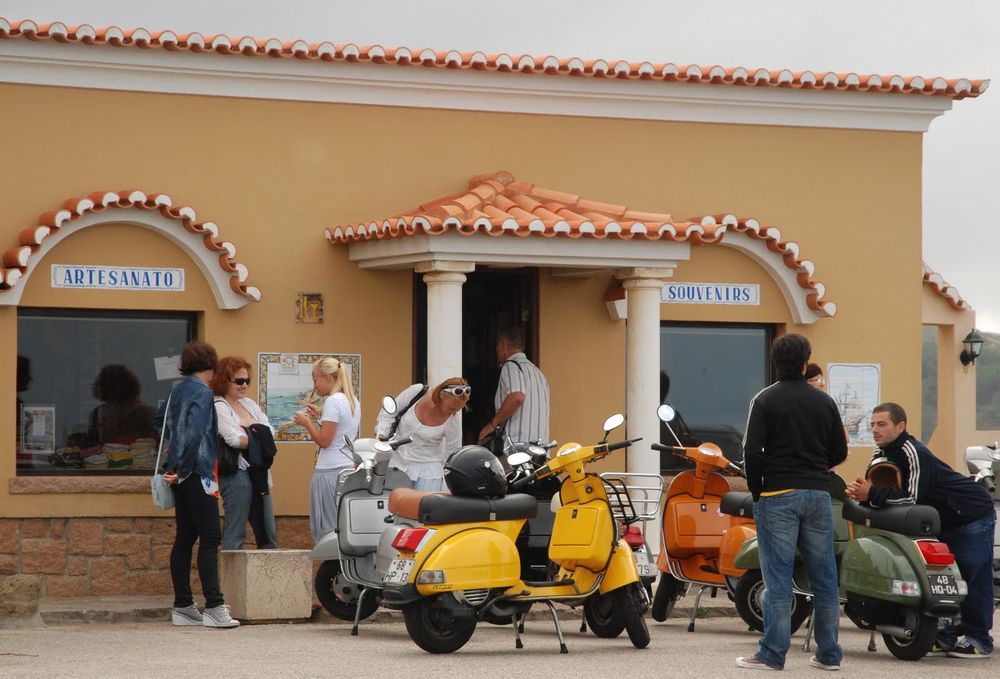 A couple of bright colored-scooters gathered in front of a light orange souvenirshop. The shop has an entrance with an overhang with roman pilars and is decorated with orange and white roof tiles. Two (friendly!) guys wearing track jackets are hanging around the scooters. A couple of women are standing in front of the shops window, probably waiting for their men to be done with buying some useless souvenirs like a knife with a handmade wooden handle or a lighter with the flag of the country, which I will NOT reveal, on it (got your hopes up, didn't I?). Although both could be usefull actually, God I really need to stop typing now. Sorry.
