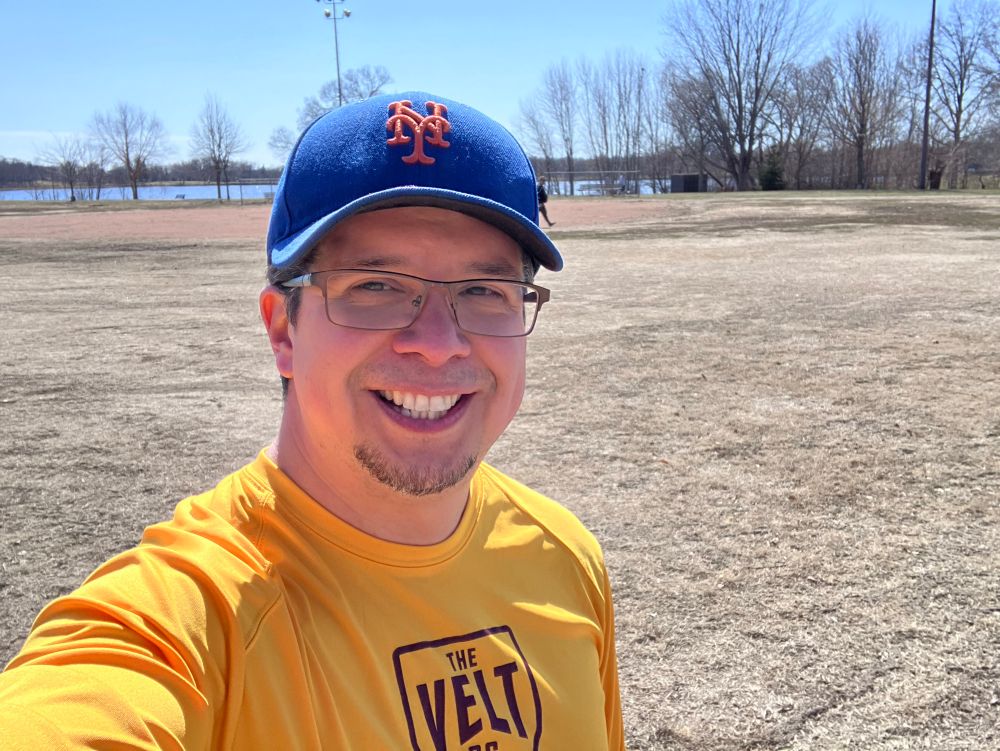 Me wearing a Mets cap and yellow shirt out at the park during a neighborhood pickup baseball game.