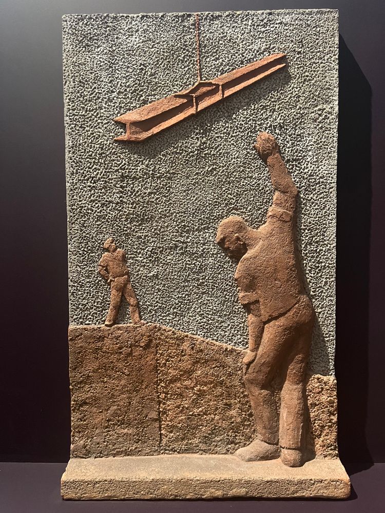 Art: A steel and concrete relief of two workmen supervising an L-beam metal girder being lowered. Earth tones. Various textures.