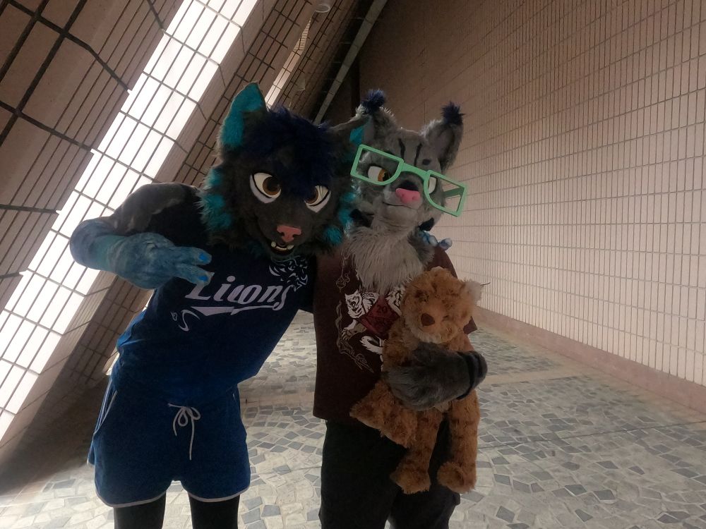 Two fursuiters, a lynx and a bobcat, posing next to a slanted architectural feature with sunlight streaming in