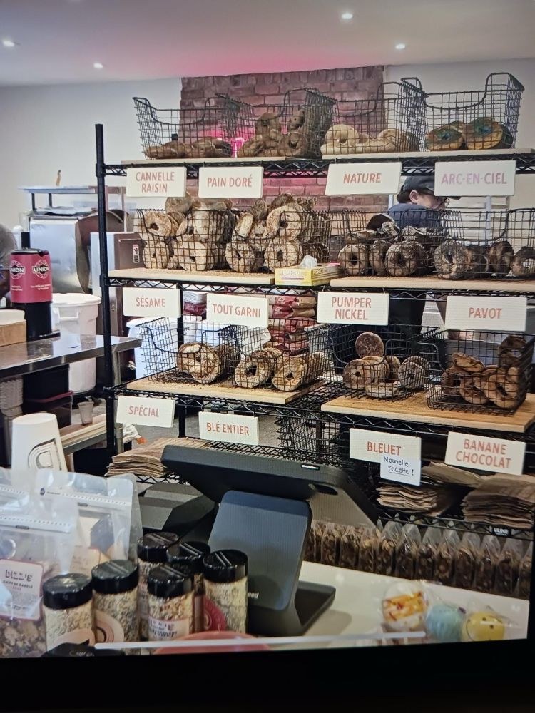 In the bagel shop Dizz‘s in Pointe Claire, a shelf with a variety of bagels in the back. On the counter several condiments…