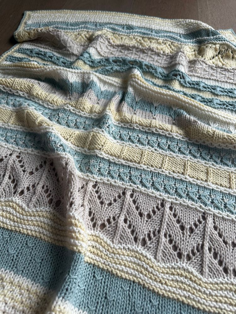 A hand knit baby blanket with various textures. Each one is a different color, varying between cream/white, soft yellow,  beige and a grey-ish teal. 