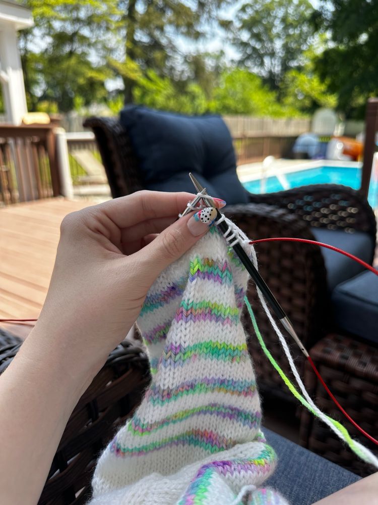 A hand holding a partially knit sleeve on a baby sweater. The sleeve is white with rainbow stripes in a variegated yarn in purples, blues and greens. The background is a pool deck in the sunlight. The nails on the hand look amazing - they’re painted with rainbow swirls with little white polka dots 