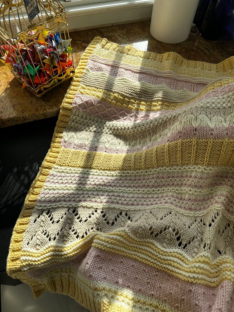 A hand knit baby blanket with various textures. Each one is a different color, varying between cream/white, soft yellow,  beige and a light pink. 