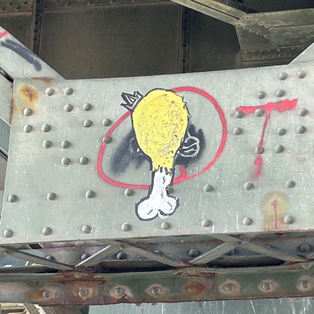 A steel support joint under a bridge, painted a dull green with rust showing through, on which has been graffitied a yellow and white cartoon fried chicken leg wearing a crown. 