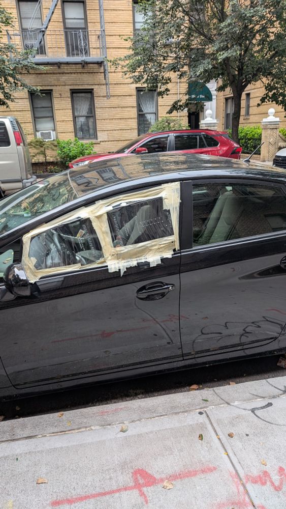 A black Toyota Prius with the front driver side window missing and clear plastic paint trays taking it's place, held there by masking tape 