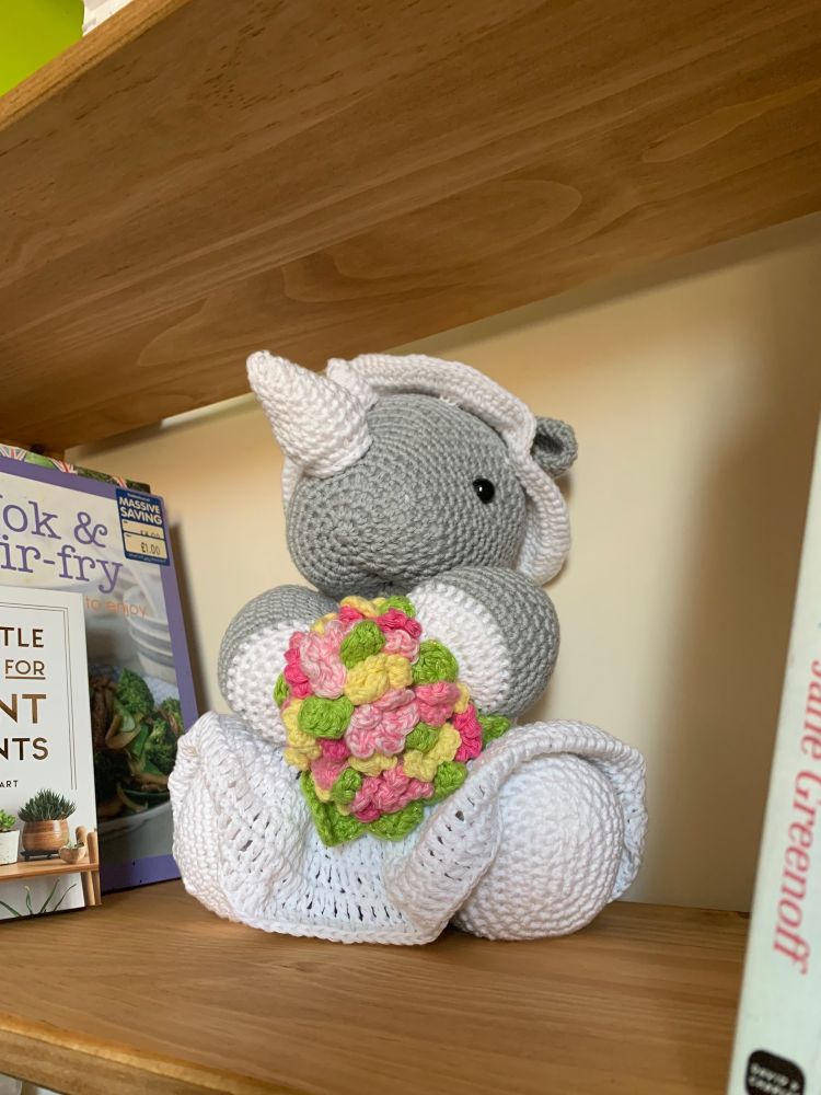 A photo of a crocheted grey rhino with white hooves and a slightly wonky white horn wearing a white dress and floppy white hat. The rhino is posed in a seated position on a pine shelf and is holding a bouquet of pink and yellow crocheted flowers with green leaves.