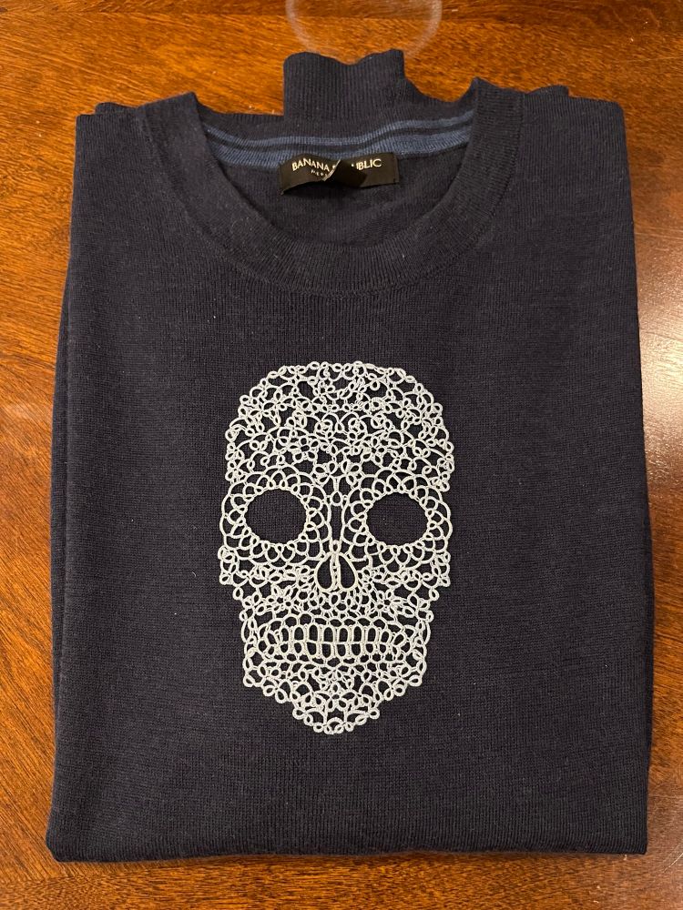 A light blue tatted skull on a dark blue lightweight wool sweater. The skull is made up of swirls and loops of thread and has the vibe of a Día de Muertos sugar skull crossed with your grandma’s pillowcase edging.