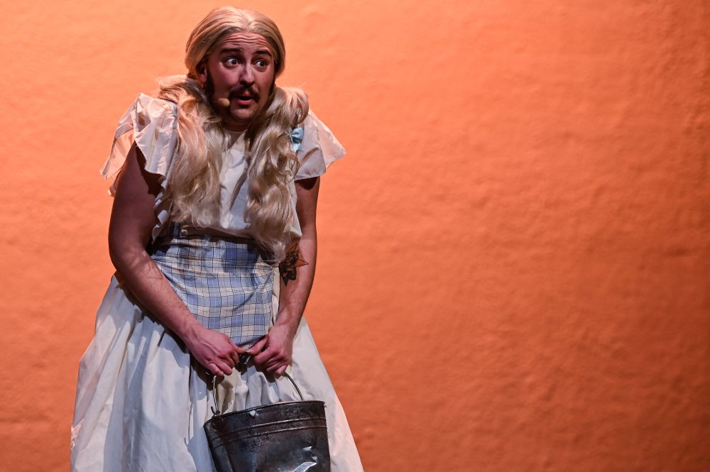 A white trans man with a brown moustache and sideburns, wearing a blonde pigtailed wig, a head mic and a gingham dress. He is clutching a silver bucket, his shoulders are raised and he has wide eyes and a cheeky expression on his face. 
