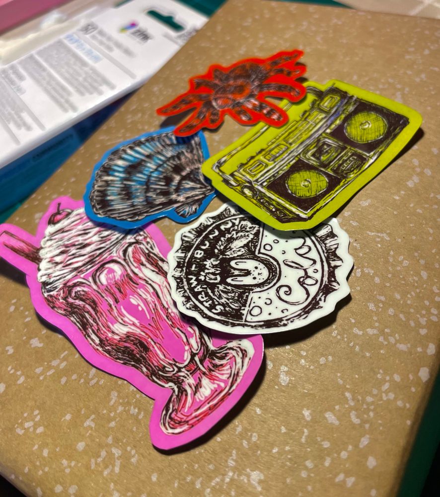 my drawings as stickers including a milkshake, boombox, pop cap, tarantula, and seashell on top of a speckled cardboard box