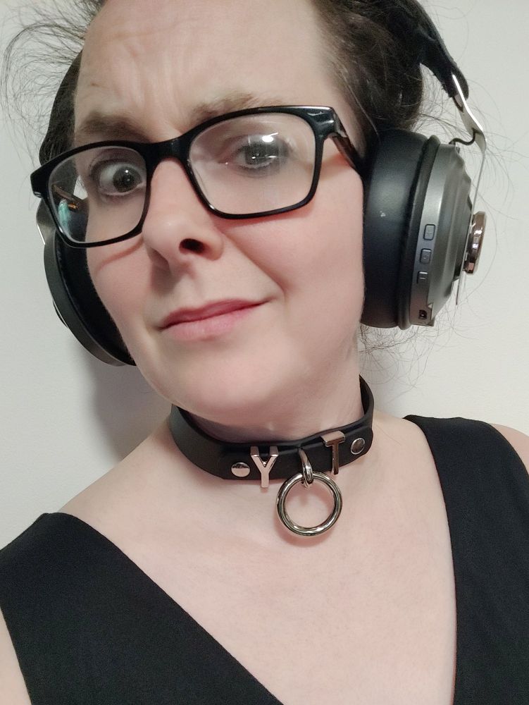 it's the maker behind Freebound - a white non binary human, kinda femme goth here. They're wearing big headphones, a puzzled expression behind nerd glasses, and a collar that says "yot". thing is, it actually says "toy", with the dangling ring forming the O - but selfies are mirrored...