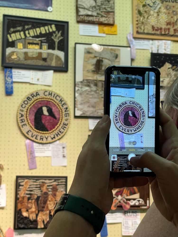 Image of a yellow peg board wall showing a piece of crop art that is round and says “Cobra Chickens Are Everywhere” with a Canada Goose head in the middle. There is a person holding up a phone taking a picture of this same piece. This is made with Black Beans, Mayocoba Beans, Red Beans, Lentils, Pumpkin Seeds and red onion skin. 