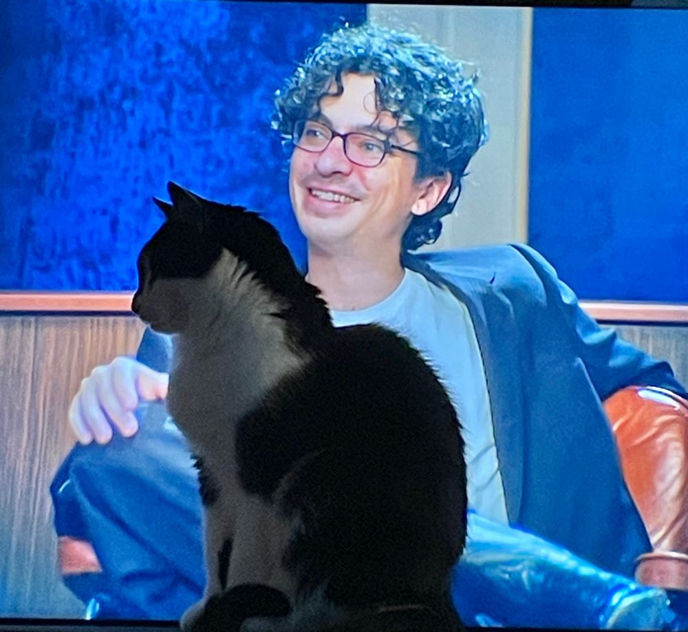 loki the tuxedo cat sitting in front of the tv while adam friedland smiles at his guest
