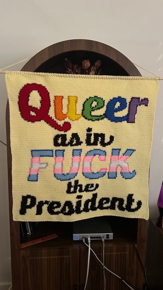 Completed Crochet tapestry that says queer as in fuck the president. Queer is done in rainbow and fuck in the trans flag. The rest of the text is black on a light yellow background. 