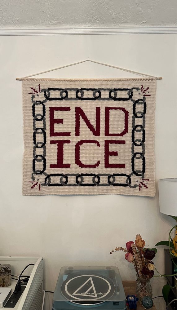 Completed crochet tapestry! It is made of cream yarn with a border of chain links made of black and grey. Each corner has a broken link. The center says END ICE in capital letters in red yarn. Tapestry hangs above my record player and vase of dried flowers 