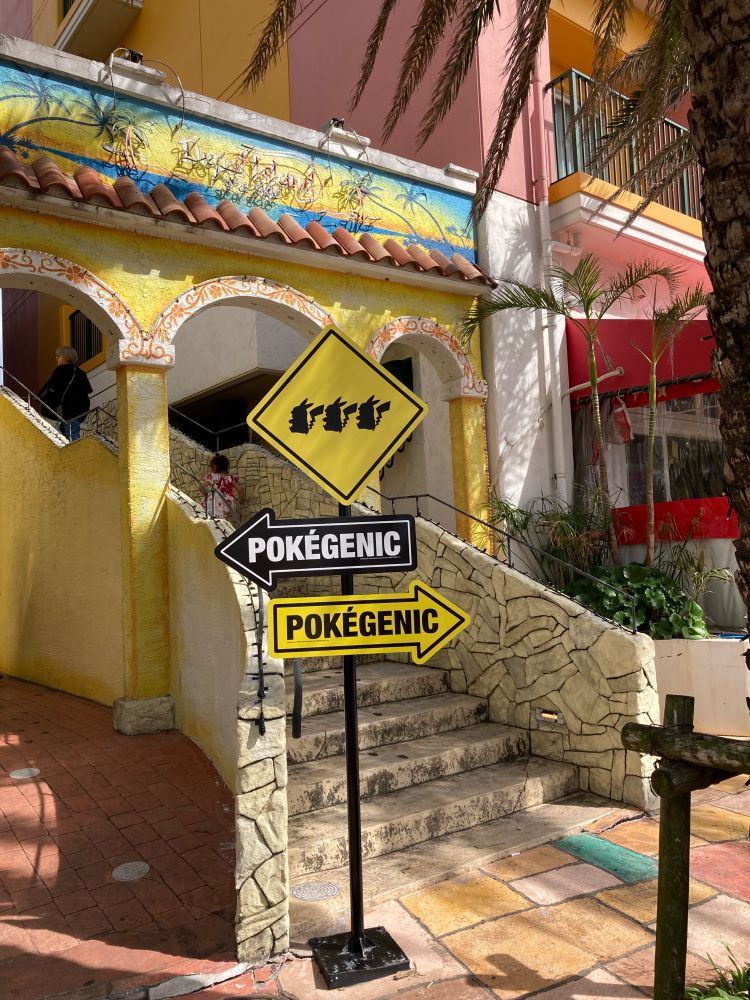 黄色、ピンク、白などに塗られたカラフルな壁に茶色系の石畳、アーチ型の入り口、ヤシの木の植栽が異国風の街並み。
写真中央の階段の脇に、3匹のピカチュウのシルエットとPOKEGENICの文字が書かれた標識が立っている。