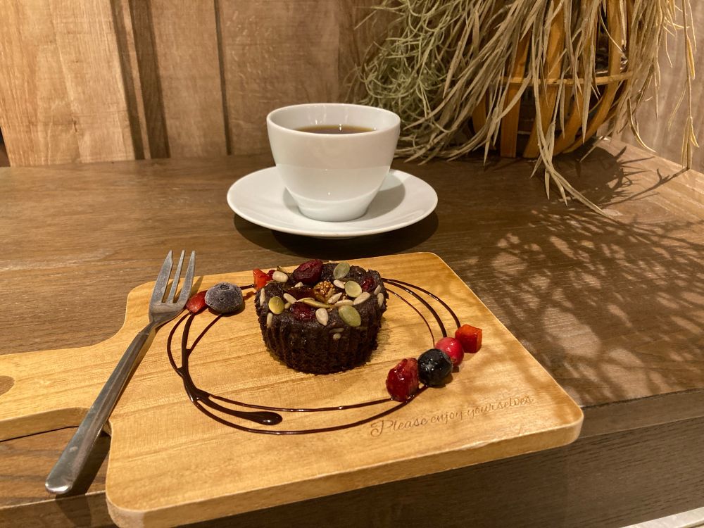 木製の机の上にチョコケーキとフォークがのった木のプレートと、奥にコーヒーの入った白いカップ＆ソーサー。
小さめの丸いケーキは上面にドライフルーツとナッツがのっていて、周囲を円を描くチョコソースとベリーで飾られている。