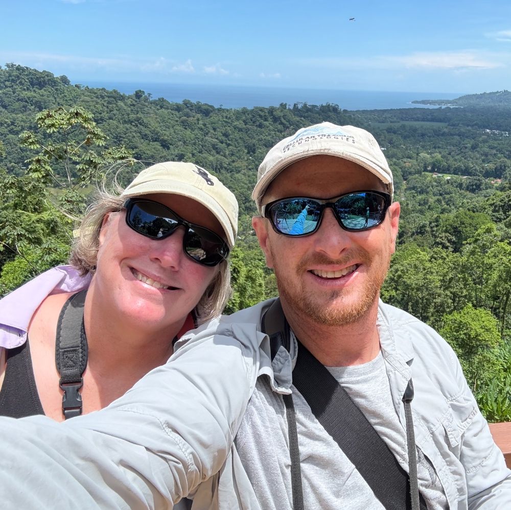 Mike Lanzone and Trish Miller smiling with a sweeping forest and coastline behind them, taken during a day of observing massive raptor migration.