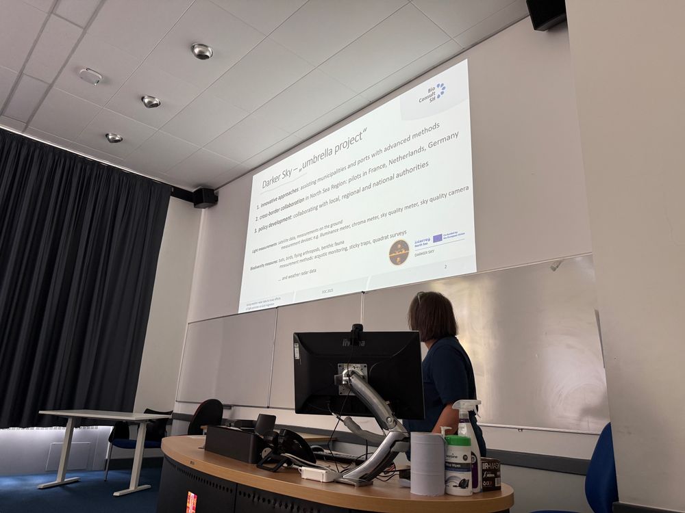 A person with short hair gives a talk at the lectern, with a slide titled “Darker Sky – umbrella project” outlining methods and cross-border collaboration to reduce light pollution.