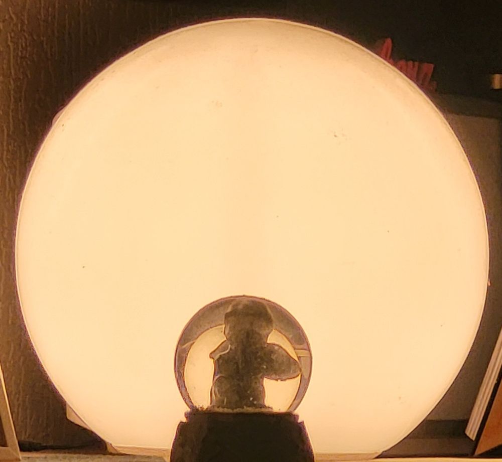 Das Bild zeigt eine Art Tischlampe mit einem großen, runden Licht diffusionskörper, der eine warme, weiche Beleuchtung bietet. Im Vordergrund befindet sich eine Schneekugel mit einem Engel, der als Schatten zu sehen ist. 

