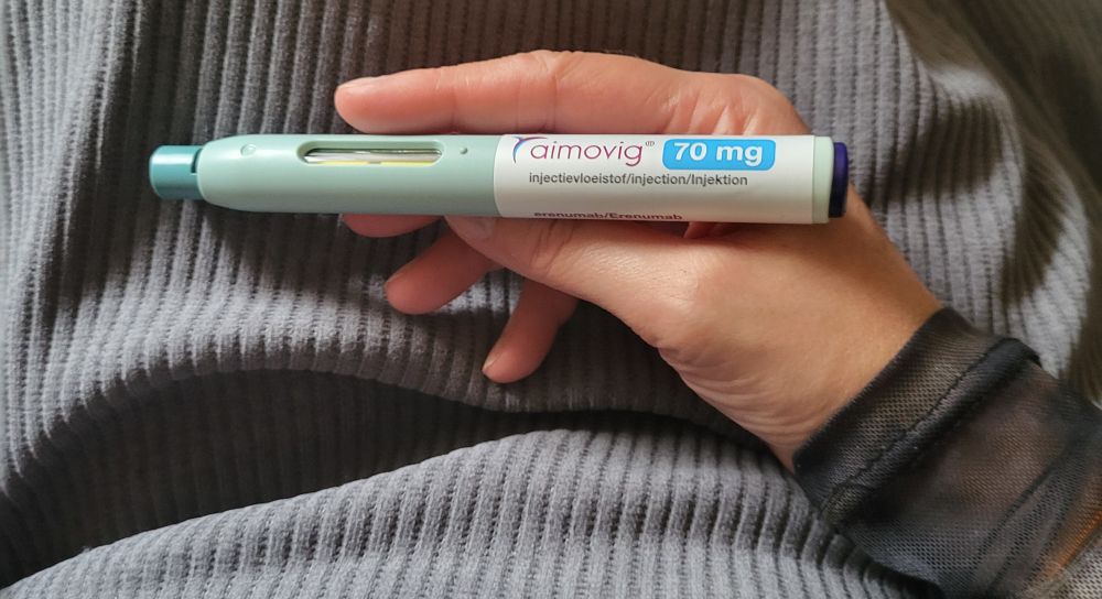 Das Bild zeigt einen Injektor (Autoinjektor) mit dem Namen "Aimovig", der eine Dosis von 70 mg enthält. Aimovig ist ein Medikament, das zur Behandlung von Migräne eingesetzt wird. 