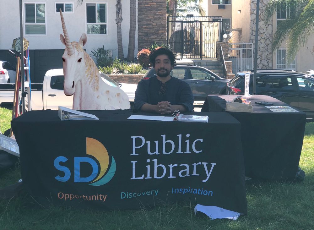 A man sitting at a booth that says San Diego Public library. I have badly photoshopped a unicorn into the chair next to him.
