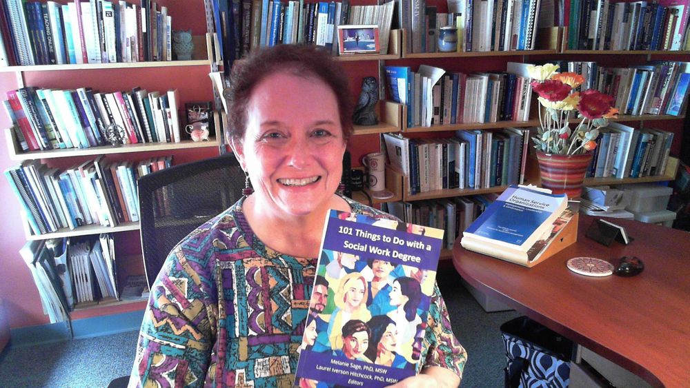Woman sitting in an office with books in background. She's  smiling and holding up the book 101 Things to Do with a Social Work Degree