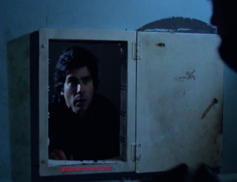 A guy looking out from another room through a hidden opening inside an open wall cabinet or fuse box.