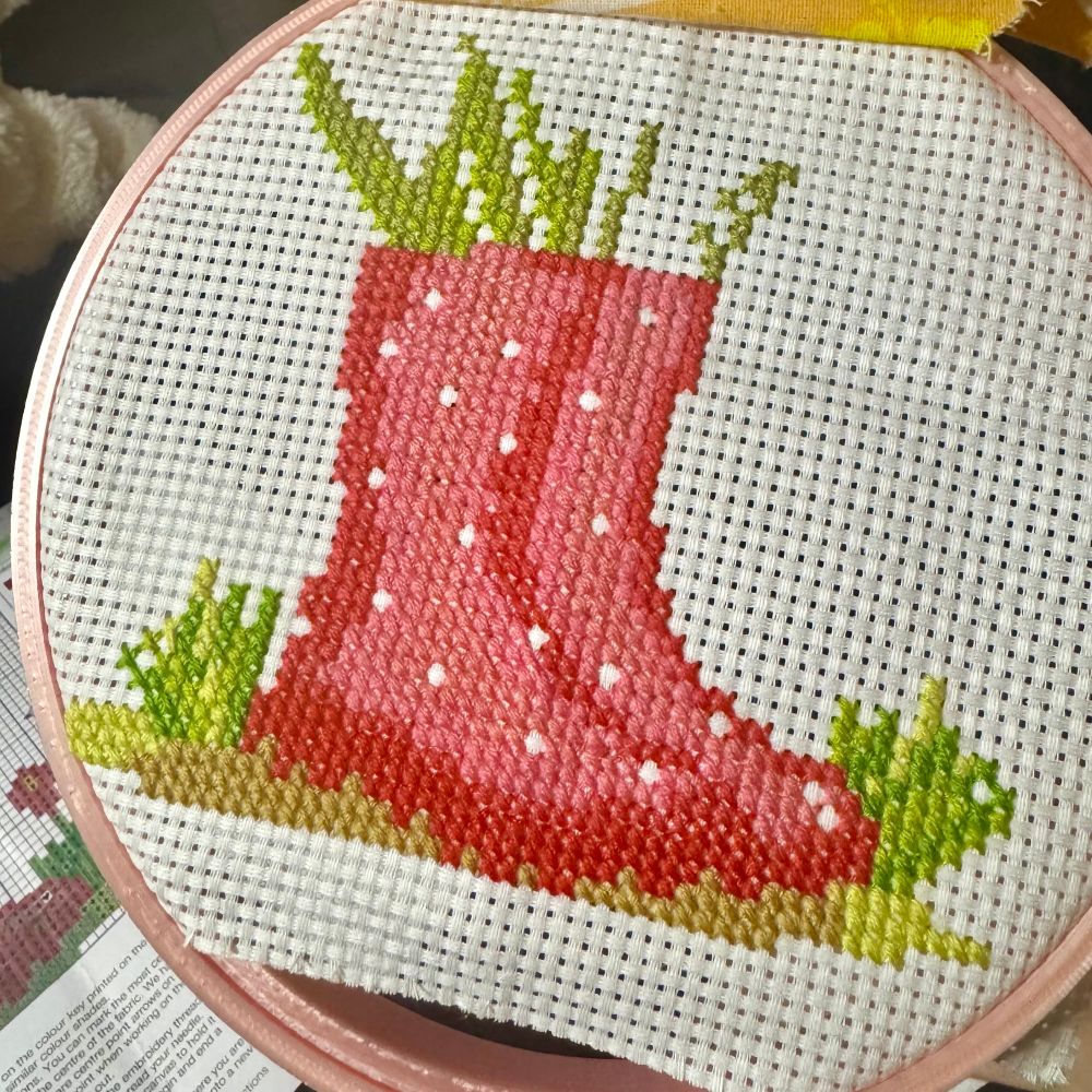 Cross-stitch work in progress on white Aida cloth showing two pink polka-dot wellington boots side-by-side, stitched in shades of pink and red with small white spots. Both boots stand on a golden-brown patch of ground with tufts of bright green grass around them. Tall green shoots are growing from the tops of both boots. Some areas of the design are still unstitched.