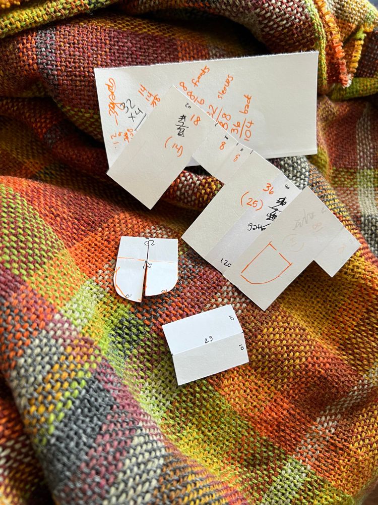 Handwoven plaid fabric with multiple pieces of paper with scribbles on them. The papers are a mini pattern for a shirt/jacket