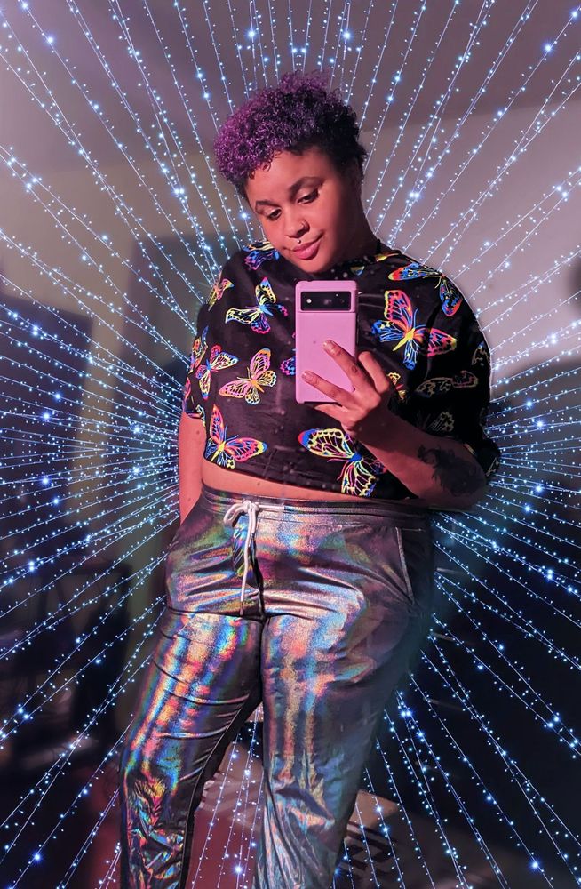 A mirror selfie from several months ago when I still had lavender hair. I'm wearing a black cropped T-shirt with printed neon butterflies on it and a pair of spectacular holographic sweatpants. I have one hand in my pocket and the other hand is holding my cell phone ,which is in a lavender case. Looking downward, my smile is slight and reminiscent of the Mona Lisa. The background has been edited with a glittery and almost starry-like backdrop to obscure the clutter in my room.