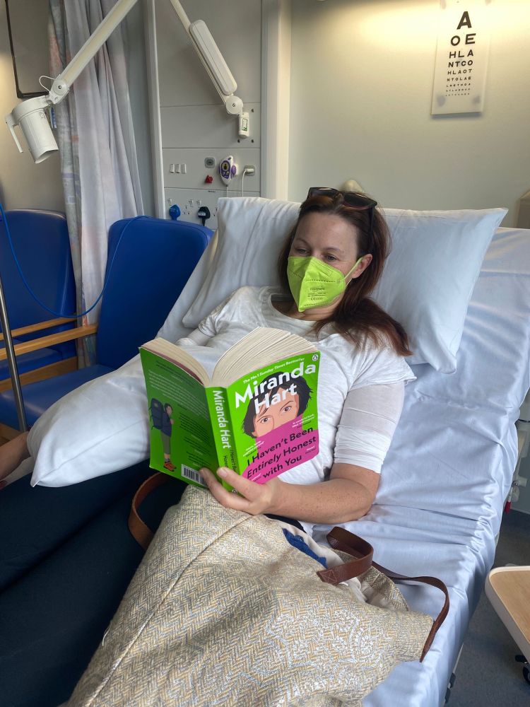 I am laying on a hospital bed in a ward at the research centre, my head is on a pillow, in the background you can see a blue chair, a wall lamp and an eye test chart on the wall.  I have long dark hair, sunglasses on my head (for some reason!) and a green face mask. I am wearing a white t-shirt and black jogging bottoms. In one hand I am holding a green book by Miranda Hard 'I haven't been entirely honest with you', the other arm is laid over a pillow and connected to the IV.
