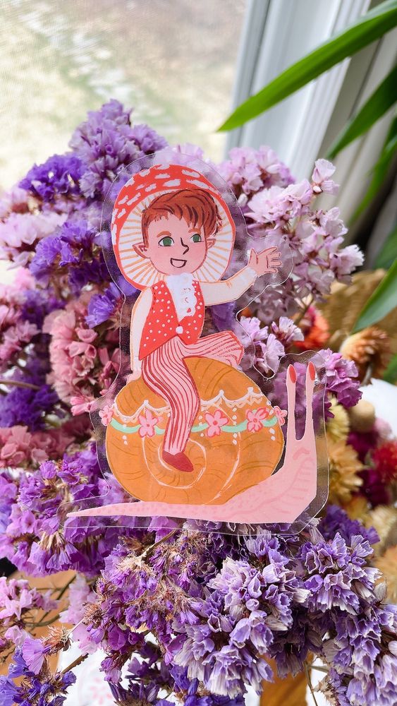 Photo of a sticker against some flowers: the sticker features a mushroom fairy waving from the back of a fancy beribboned snail.
