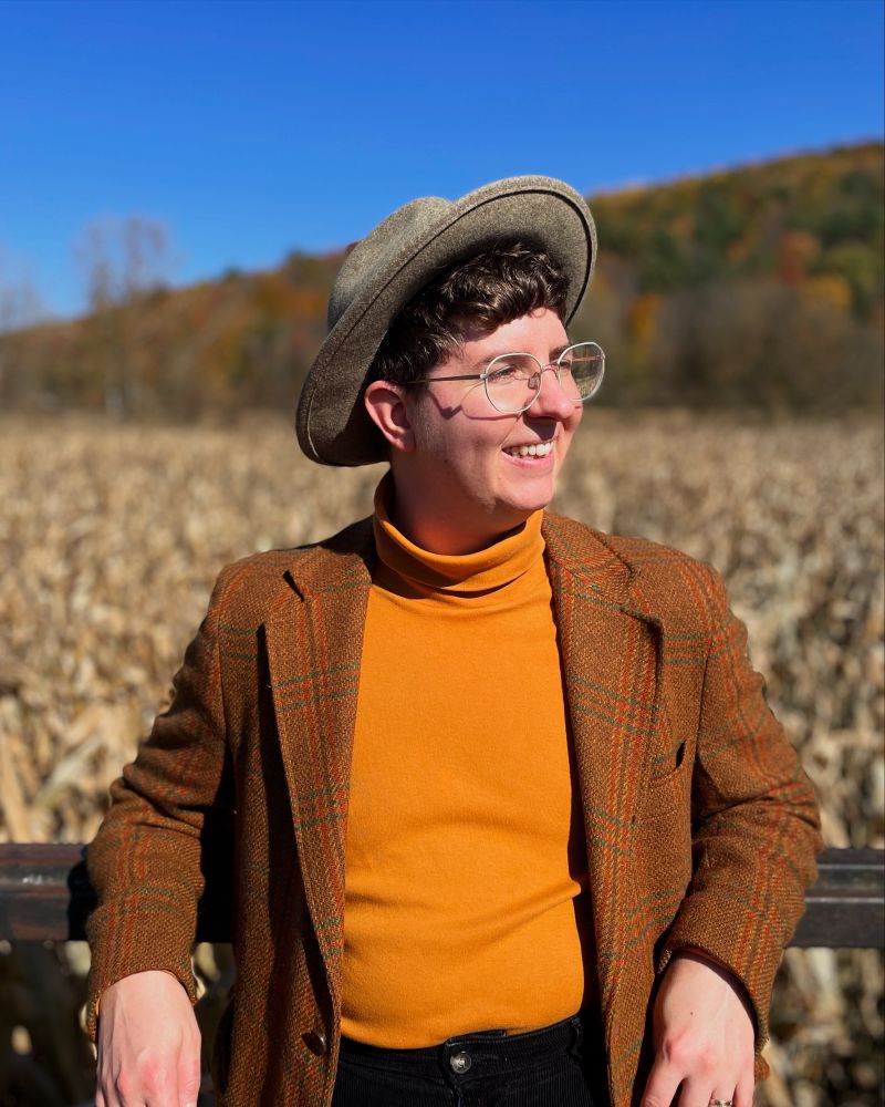 Photo of the artist: a white trans guy with fluffy brown hair, squinting into the sun with a golden cornfield stretching away behind him. He's wearing a bright golden turtleneck and a cool tweed jacket.