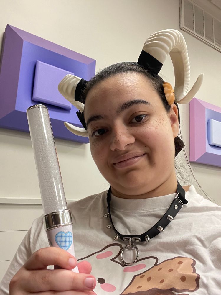 A person wearing ram horns, a spiked collar, cookie pins and a brown bow is holding a lightstick and smiling