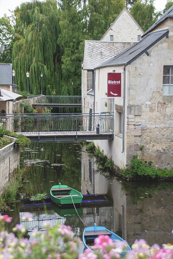 A bistro in an old building in the medieval town of Bayeux, France is framed by a row of flowers as a small boat rests in the surrounding water.