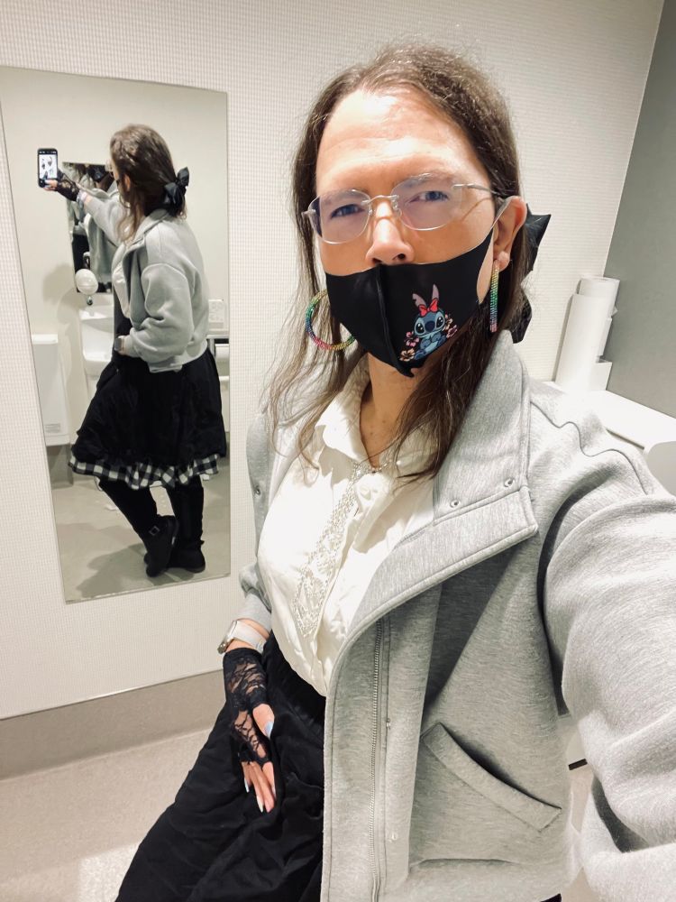 Trans woman wearing rainbow earring loops, black hair ribbon-bow 🎀, black Stitch ™️ mask, black lace gloves, trans themed nail polish, gray H&M sweater jacket, black & white tartan skirt, and a bejeweled necklace shaped like a tie.
She posing in a mirror displaying both front and back of her outfit while her jacket is open. 