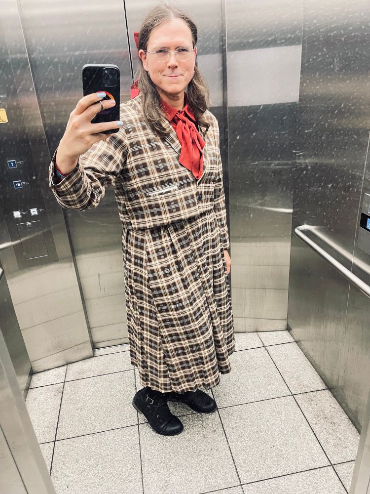 Trans woman in a fall PUNYUS outfit. Red blouse with ribbon and red hair bow. Black boots. Smiling into elevator mirror. 