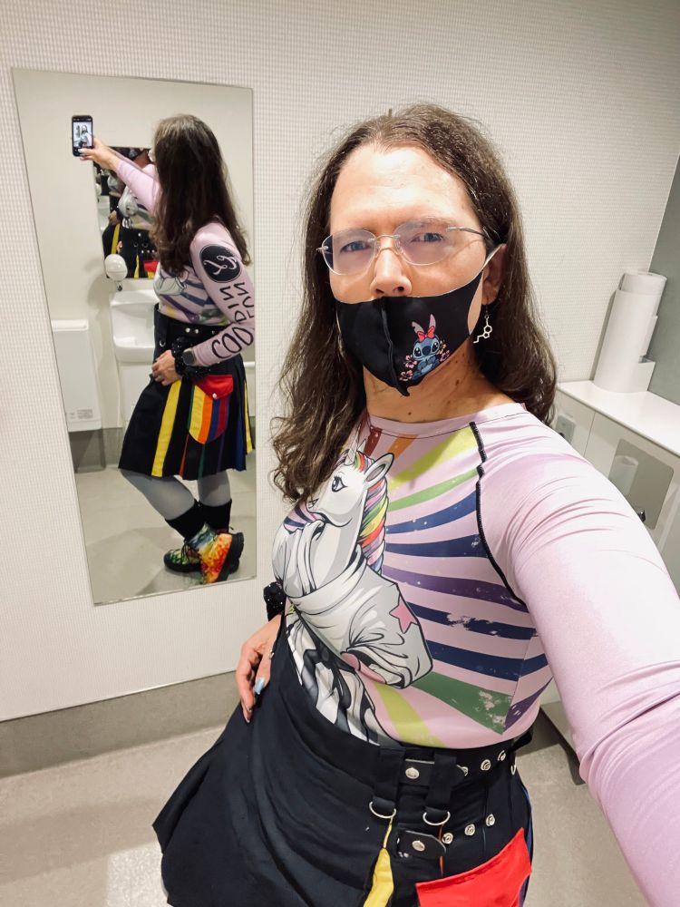 Trans woman wearing glasses, Stitch ™️Mask, estrogen symbol and transgender earrings. Her long hair drapes over her rainbow 🌈 unicorn in a kendo pose shirt. Nails 💅 painted with trans 🏳️‍⚧️ colors. Rainbow kilt and sky-blue pantyhose. Thick black socks and custom made “Vegan Fall Pride” boots. She’s using the front facing camera with mirror to capture full body image. 