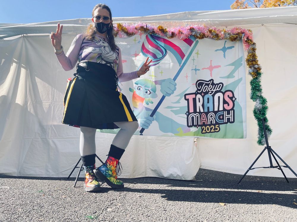 Trans woman wearing glasses, Stitch ™️Mask, estrogen symbol and transgender earrings. Her long hair drapes over her rainbow 🌈 unicorn in a kendo pose shirt. Nails 💅 painted with trans 🏳️‍⚧️ colors. Rainbow kilt and sky-blue pantyhose. Thick black socks and custom made “Vegan Fall Pride” boots. 
