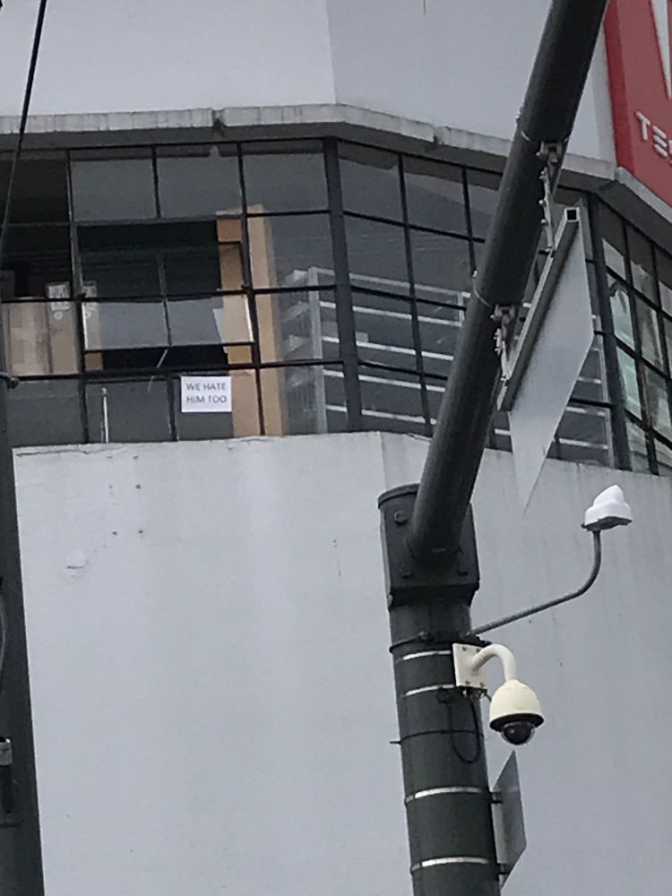 A tiny handwritten sign at the Tesla dealership as a big crowd protests Musk reads “We hate him too” yup 👍 